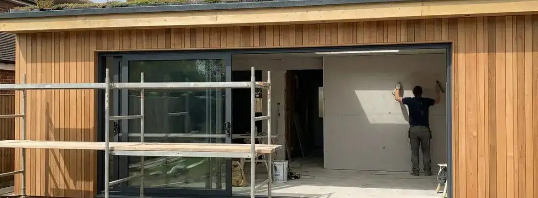 Un ampliamento in costruzione: rivestimento in legno, vetrata scorrevole, ponteggio e un operaio che monta il cartongesso.